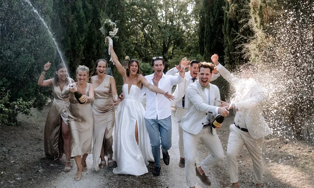 Feierlicher Moment voller Energie: Braut und Bräutigam mitten im Freundeskreis bei einer spritzigen Sektdusche im Grünen.