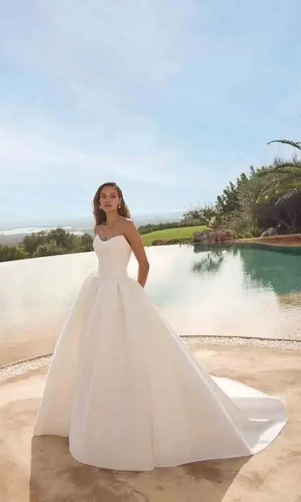 Minimalistisches Brautkleid mit weitem Rock und Schleppe, getragen von einer Braut am sonnigen Infinity-Pool.