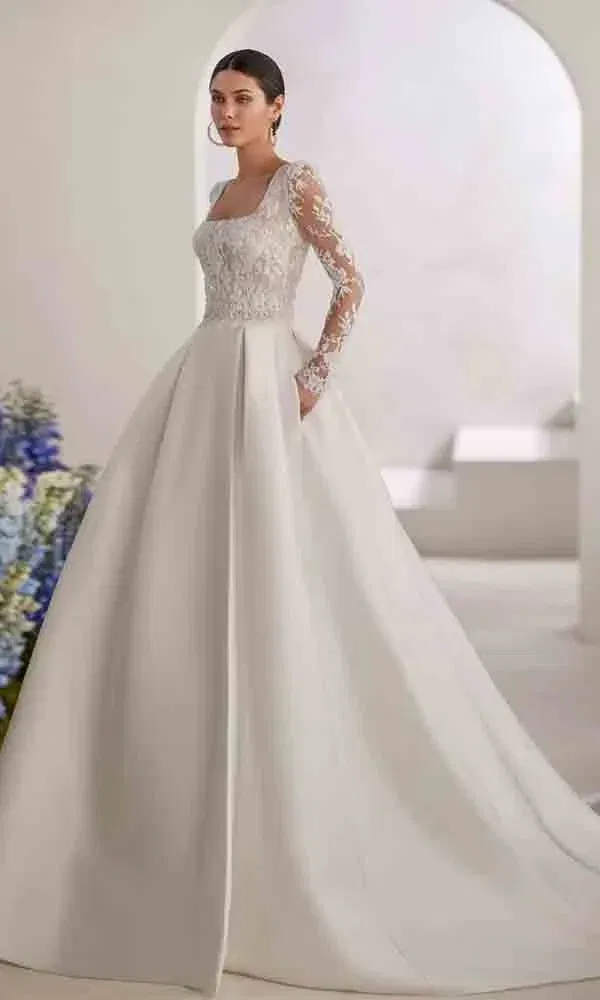 Zeitloser Brautlook mit aufwendig besticktem Oberteil, langen Spitzenärmeln und weitem Rock in heller Kulisse.