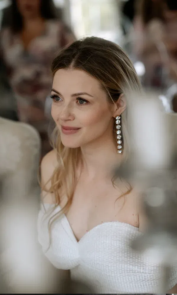 Nahaufnahme einer nachdenklich blickenden Braut in natürlichem Licht, im Hintergrund verschwommen Hochzeitsgäste.