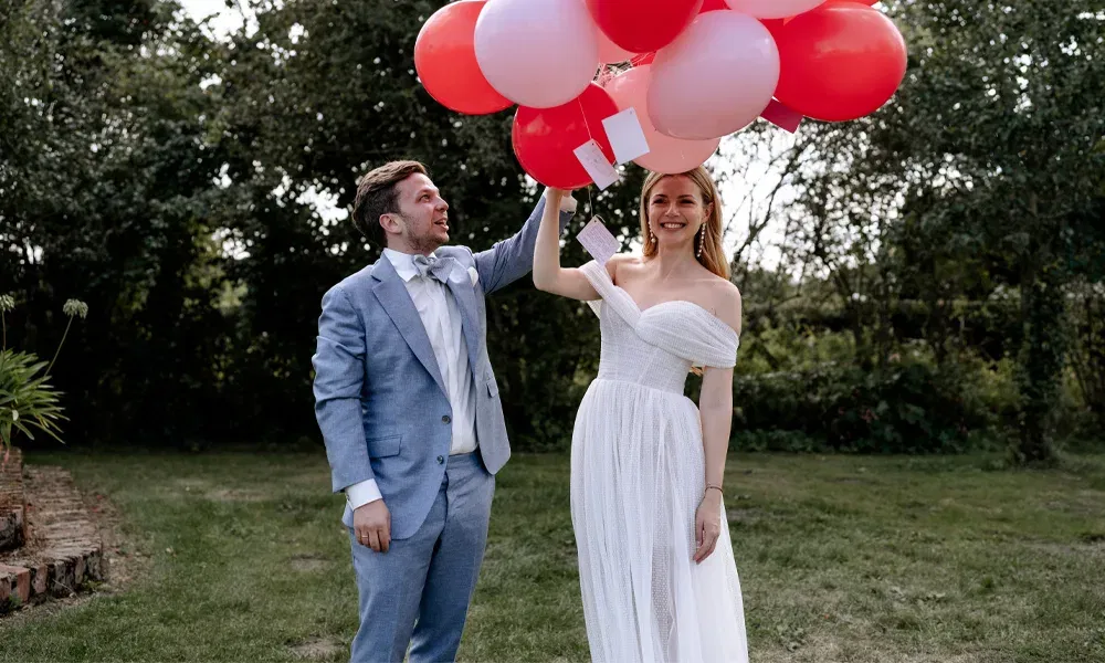 Im Garten hält die Braut rote und rosa Luftballons mit angehängten Karten, während das Paar diesen Moment lachend genießt.