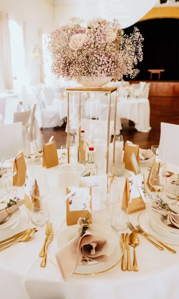 Tischdekoration mit opulenter Blumensäule, goldenen Akzenten, Geschenkpapier-Tüten und Namenskärtchen zur Hochzeit.