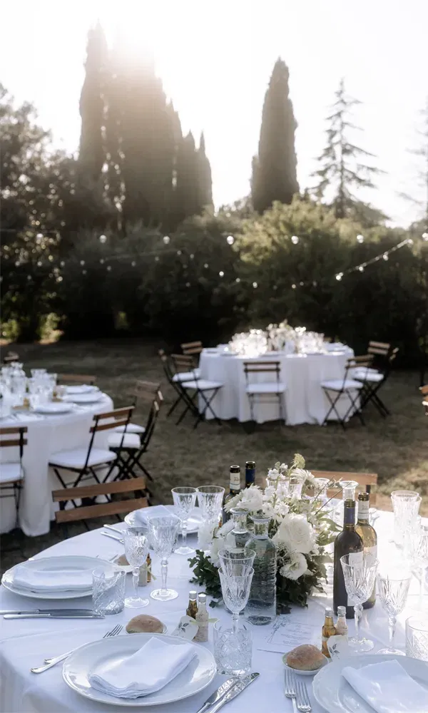 Romantisch gedeckte Tische auf einer Gartenhochzeit – weiße Tischdecken, Gläser, Brot und mediterrane Details im Sonnenlicht.