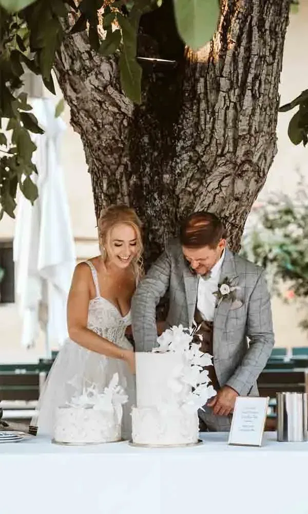 Das Brautpaar lacht beim Anschneiden der kunstvoll dekorierten Hochzeitstorte unter freiem Himmel.