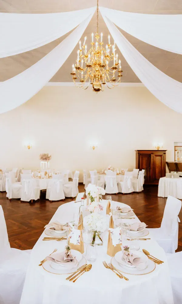 Langgedeckter Hochzeitstisch mit floraler Mitte, goldfarbenem Besteck und festlichem Ambiente unter einem prunkvollen Kronleuchter.