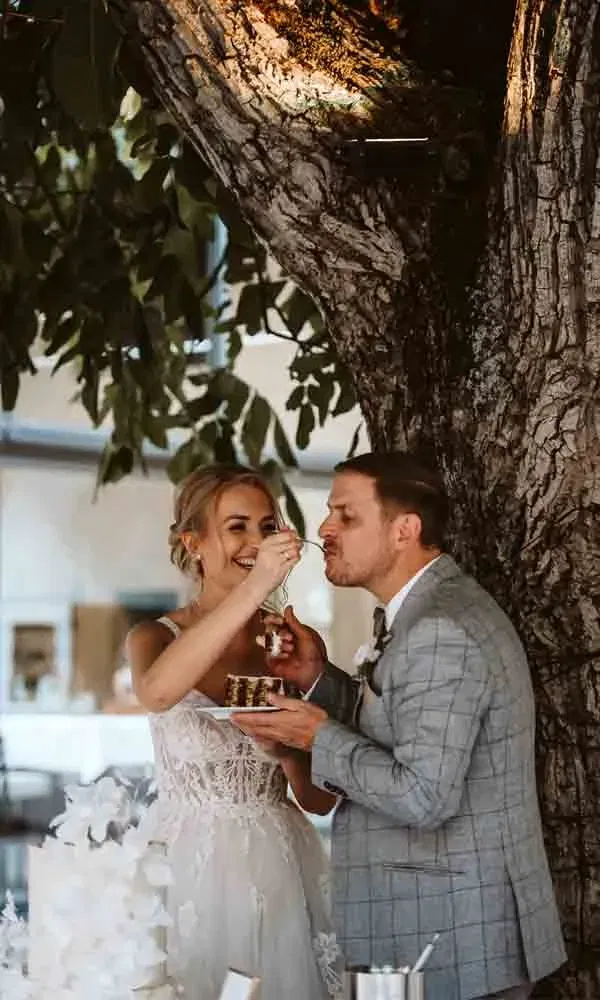 Braut und Bräutigam beim gegenseitigen Torte-Füttern unter einem Baum – ein verspielter Hochzeitsschnappschuss.