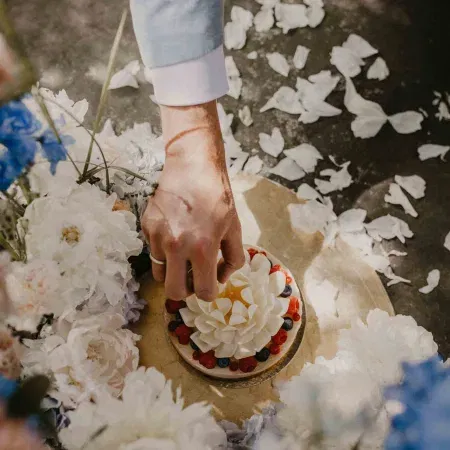 Stilvolles Hochzeitstorten-Detail: Bräutigamhand greift nach fruchtiger Torte mit Blütenoptik im Blütenmeer