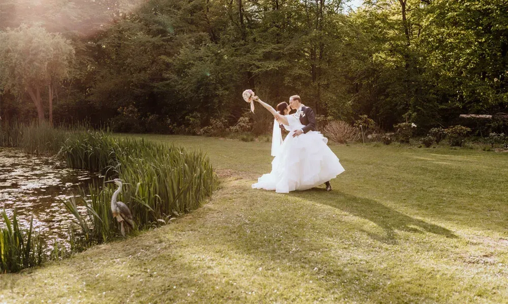 Weitwinkelaufnahme eines Hochzeitspaares im Grünen, die Braut streckt den Blumenstrauß hoch – im Vordergrund ein Reiher am Wasser.