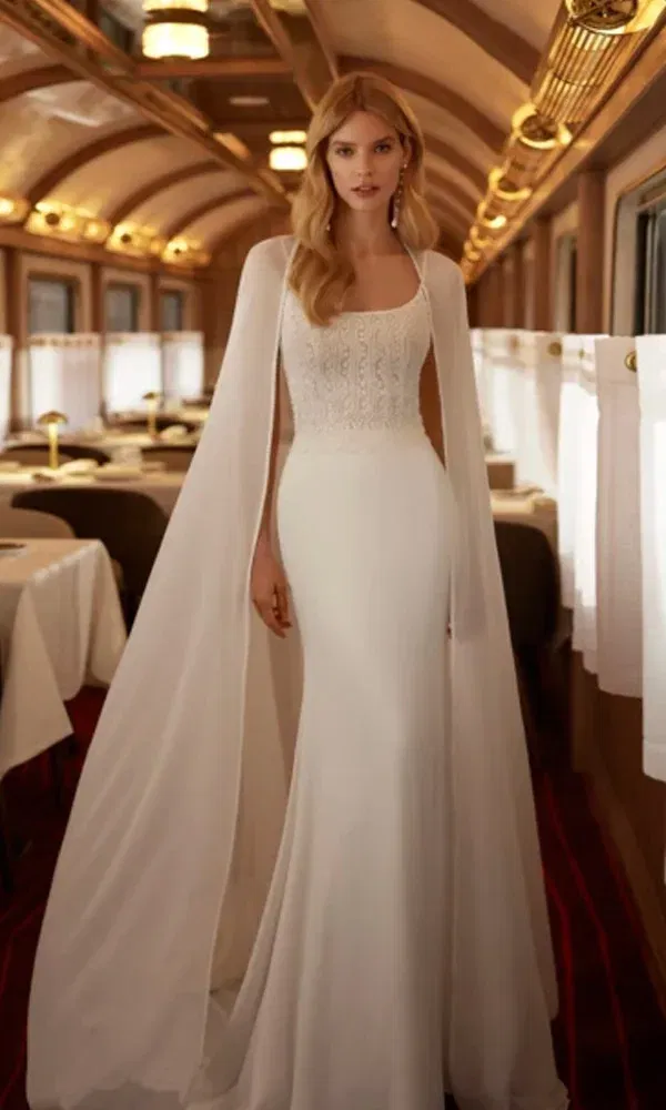 Figurbetontes Brautkleid mit filigraner Stickerei am Oberteil und bodenlangem Cape, aufgenommen in einem nostalgisch dekorierten Waggon.