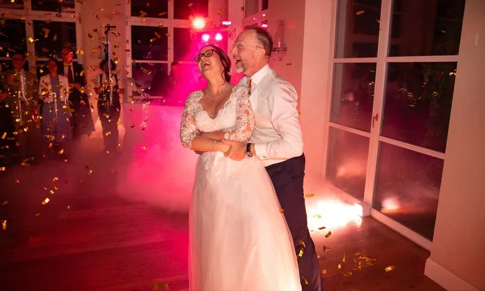 Braut und Bräutigam tanzen lachend im goldenen Konfettiregen, umgeben von rosa Licht und Nebel.