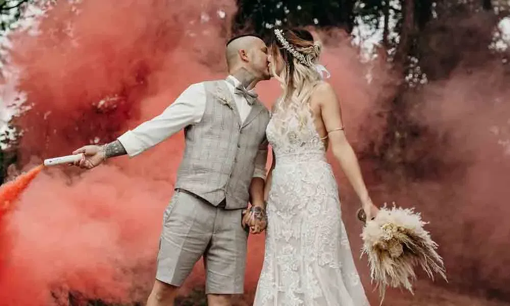 Boho-Hochzeit mit Wow-Effekt: Braut in Spitzenkleid und Bräutigam in Shorts posieren mit Smoke Bomb in rotem Nebel.