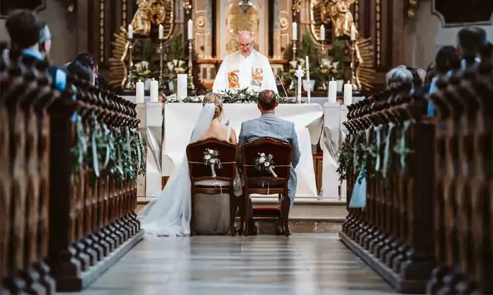 Feierliche Hochzeitsszene in der Kirche: Das Paar sitzt vor dem Priester, der Traualtar ist festlich dekoriert.