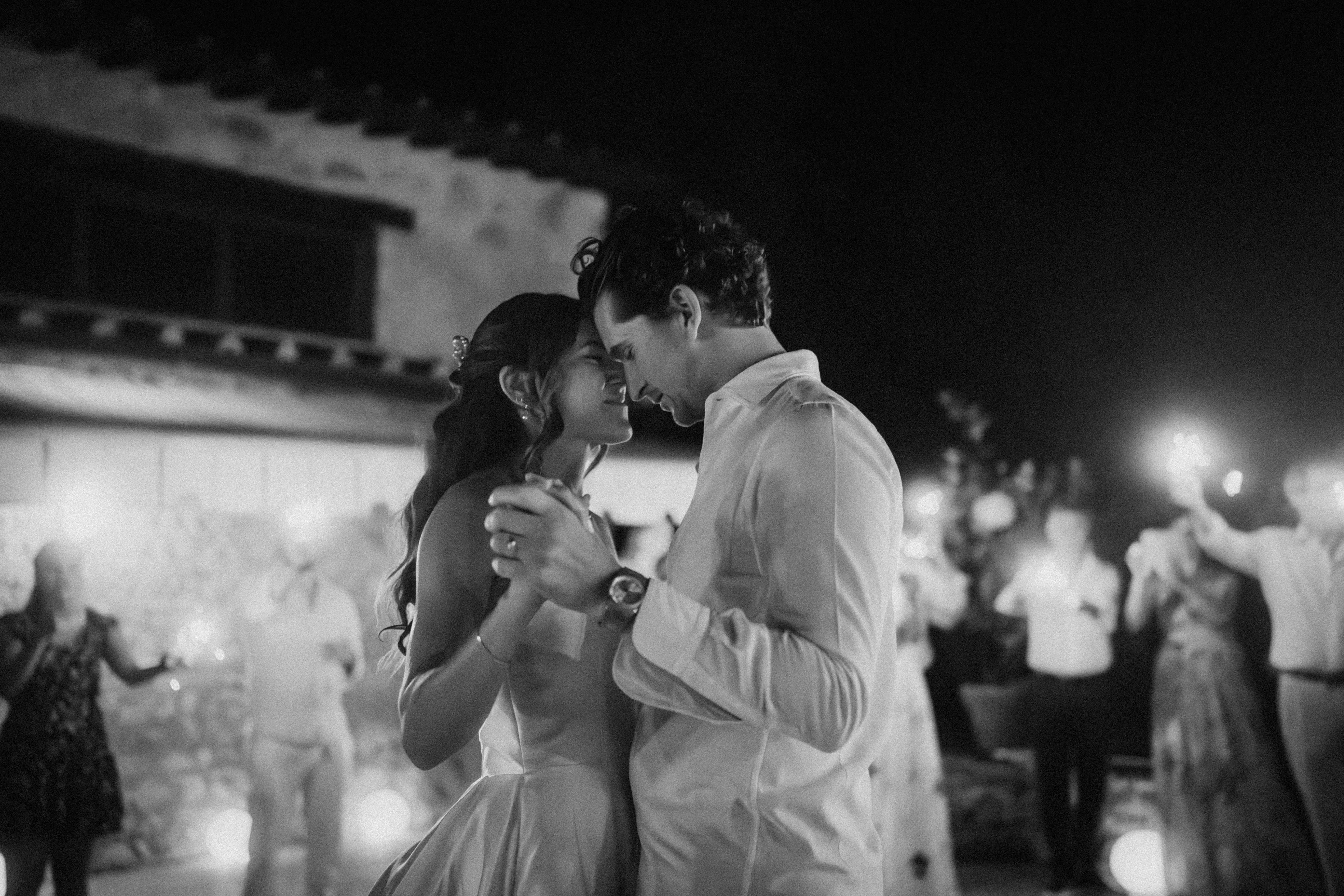 Erster Tanz des Brautpaars bei stimmungsvoller Abendfeier – emotionale Szene in Schwarz-Weiß mit leuchtenden Gästen im Hintergrund.