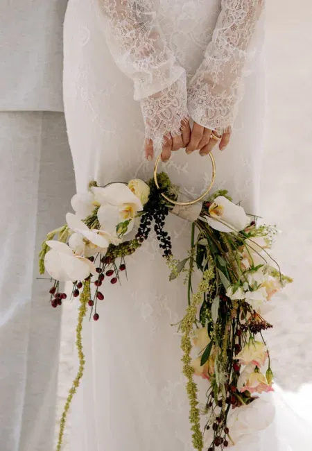 Braut in Spitzenkleid hält runden Blumenring mit hängenden Blüten, Orchideen und wilden Zweigen