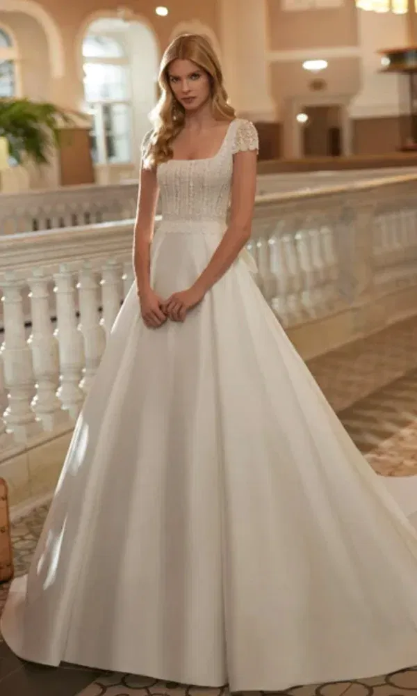 Romantische Robe mit strukturiertem Spitzenmieder und Cap-Ärmeln, präsentiert von einer Braut in einem lichtdurchfluteten Festsaal.