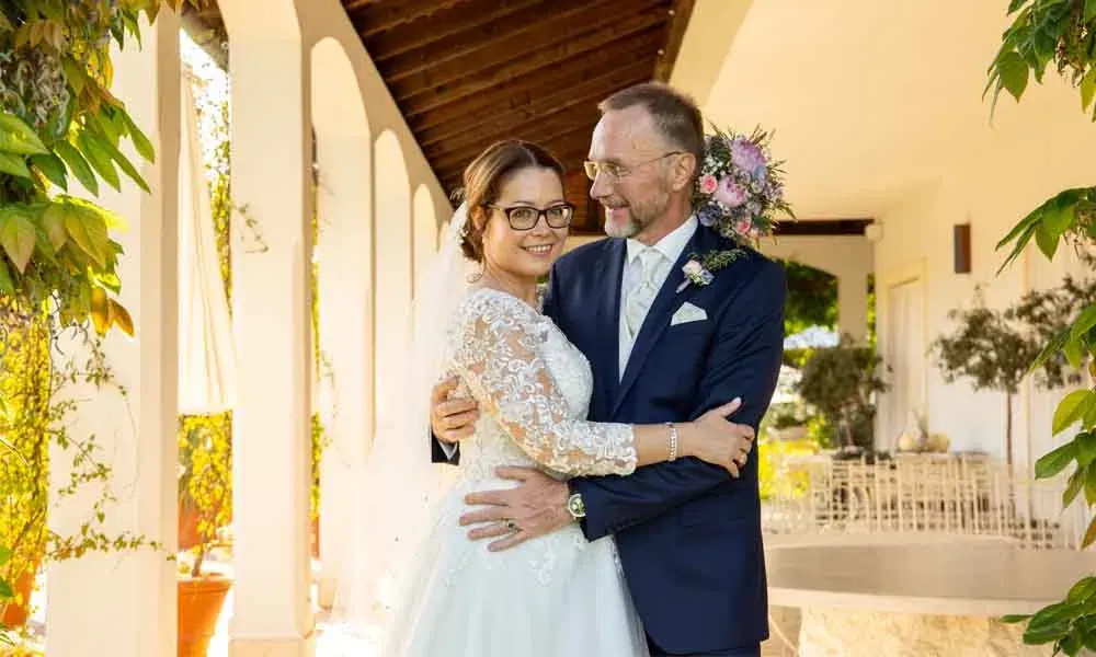 Braut und Bräutigam umarmen sich glücklich im Arkadengang ihrer Hochzeitslocation, sie tragen elegante Festkleidung.