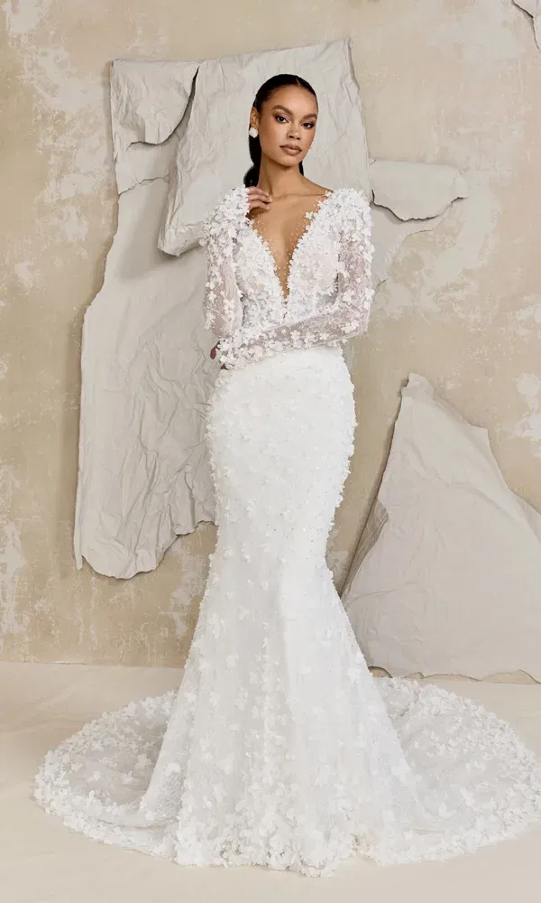 Elegante Braut in einem modernen Spitzenkleid mit dreidimensionalen Blüten und transparenter Oberpartie, fotografiert vor einer kunstvoll zerknitterten Papierwand in neutralen Tönen.