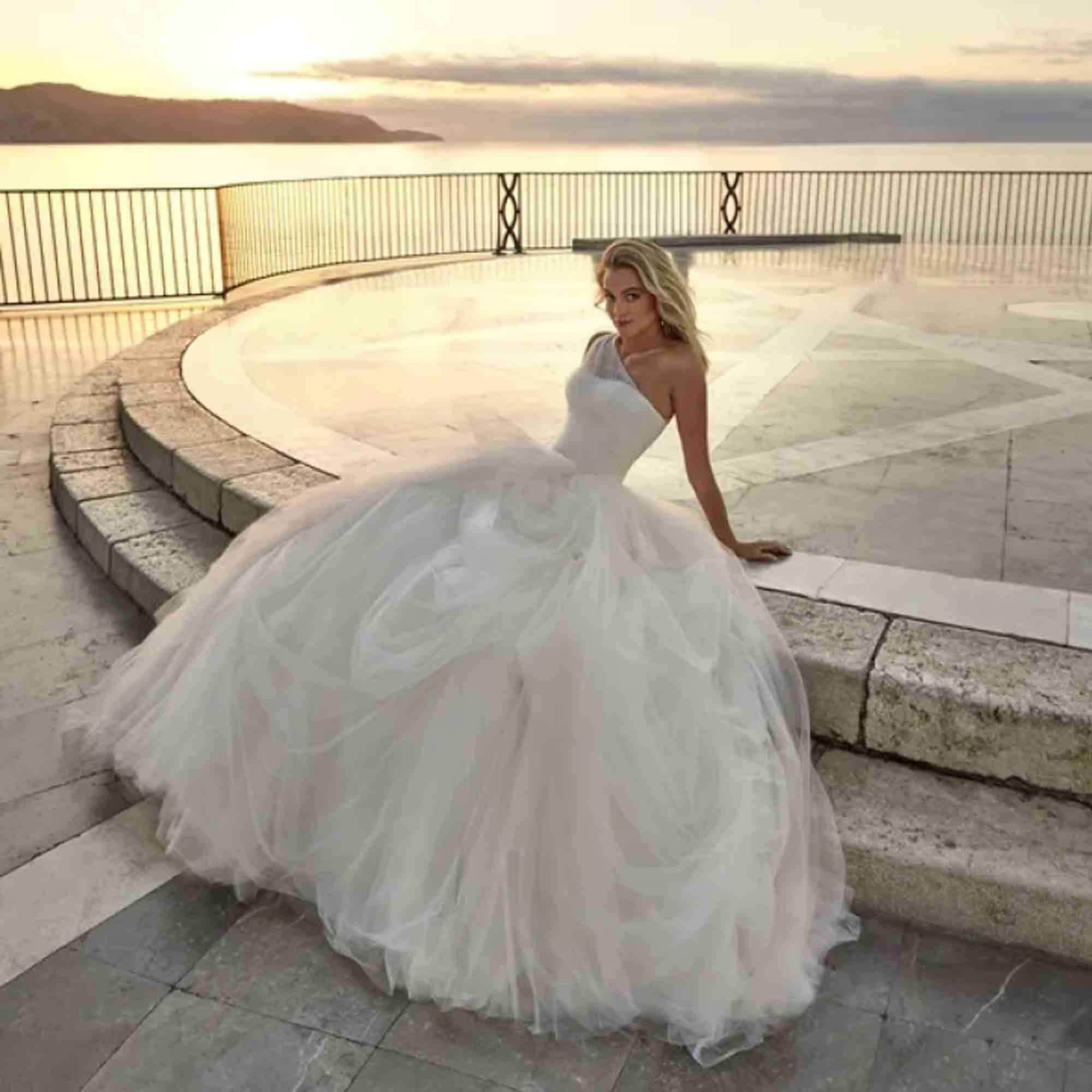 Elegante Frau im glamourösen One-Shoulder-Brautkleid mit ausladendem Rock sitzt entspannt an einer steinernen Balustrade mit Blick aufs Meer im Sonnenuntergang.