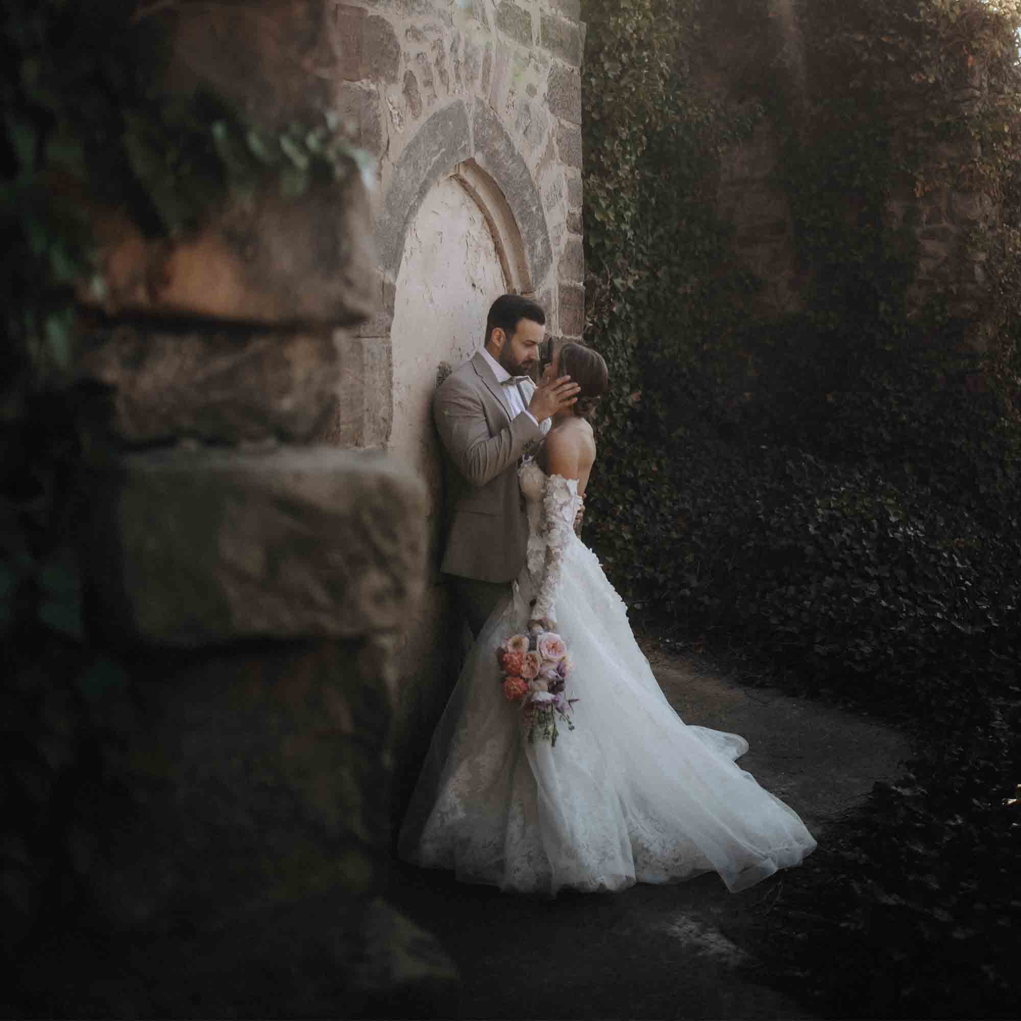 Hochzeitspaar steht innig an einer alten Steinmauer, die Braut im Spitzenkleid hält einen Blumenstrauß mit pastellfarbenen Rosen.