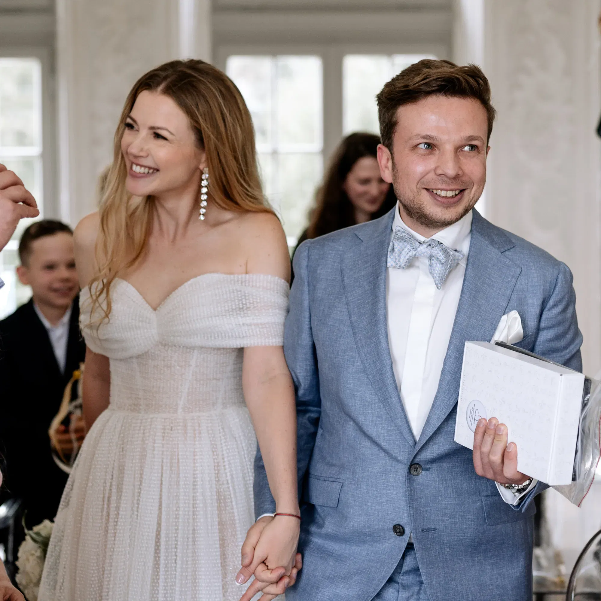 Hochzeitsmoment voller Freude: Die Braut in einem eleganten Off-Shoulder-Kleid und der Bräutigam im hellblauen Anzug lächeln in die Runde.