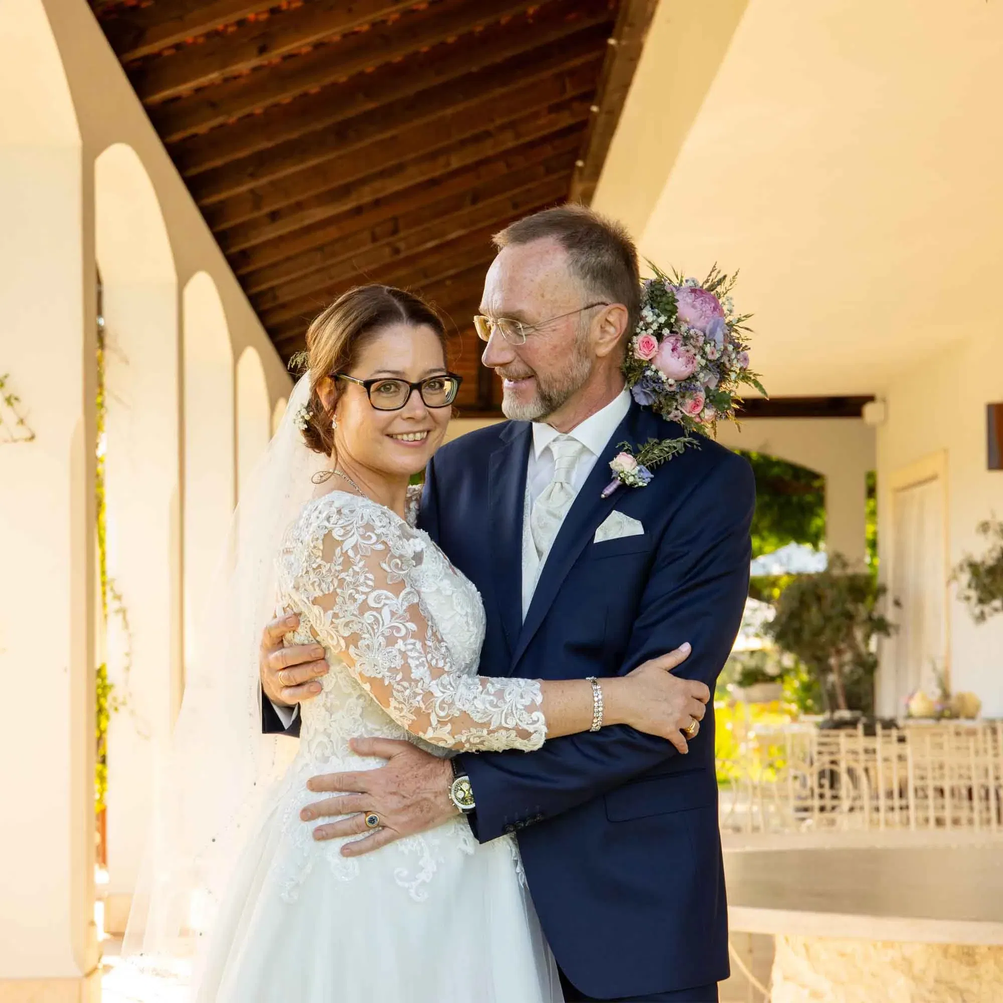 Braut und Bräutigam umarmen sich lächelnd unter einem Arkadengang, beide in festlicher Hochzeitskleidung.