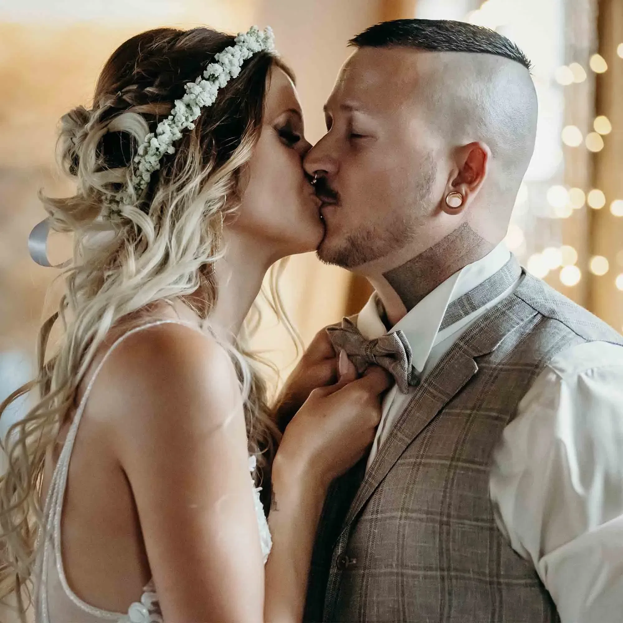 Zärtlicher Hochzeitsmoment: Braut im Boho-Stil zieht den Bräutigam zum Kuss an sich, während Lichter im Hintergrund leuchten.