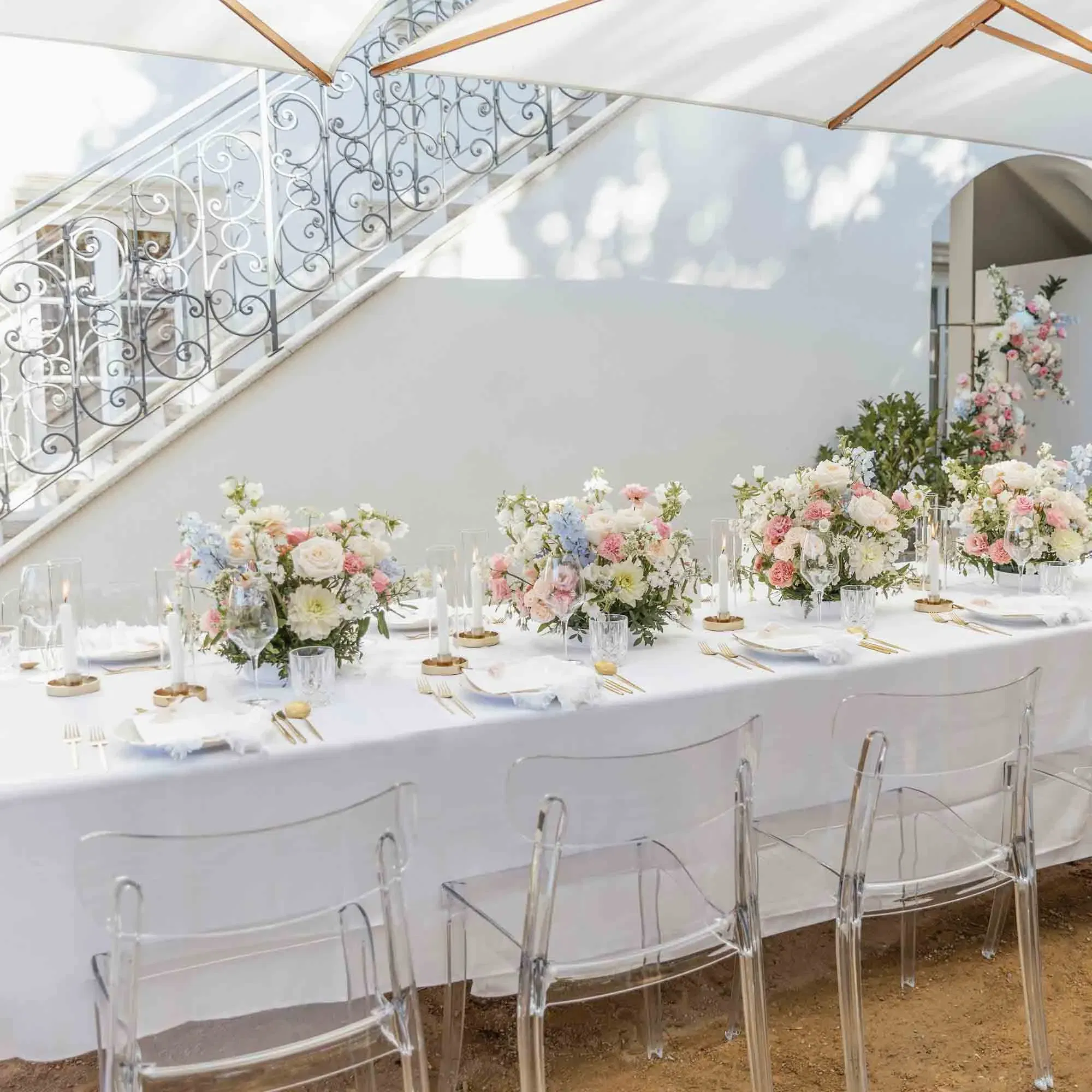 Elegante Hochzeitstischdekoration im Garten, mit pastellfarbenen Blüten, minimalistischen Gläsern und modernen Stühlen aus Acryl.