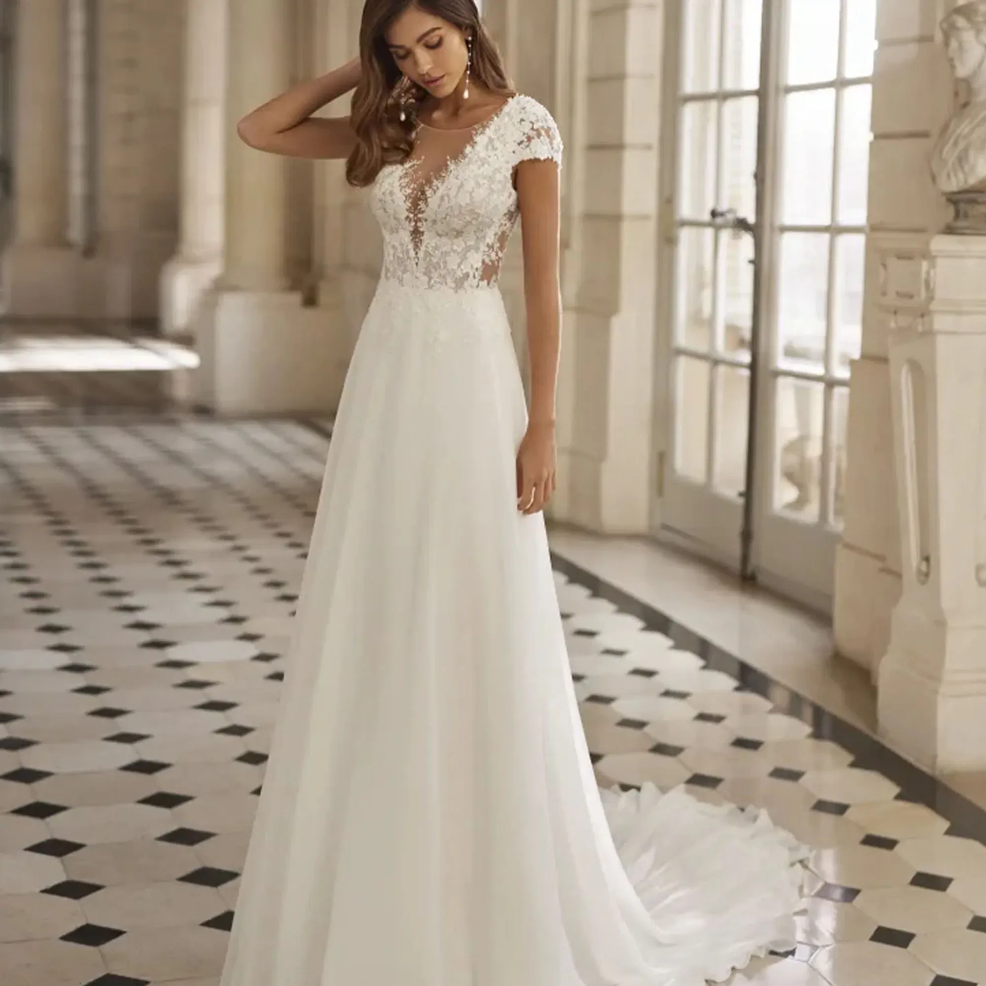 Moderne Braut in einem fließenden Kleid mit Cap-Ärmeln, tiefem V-Ausschnitt und feinen Spitzenapplikationen posiert in einem lichtdurchfluteten Schlossambiente.