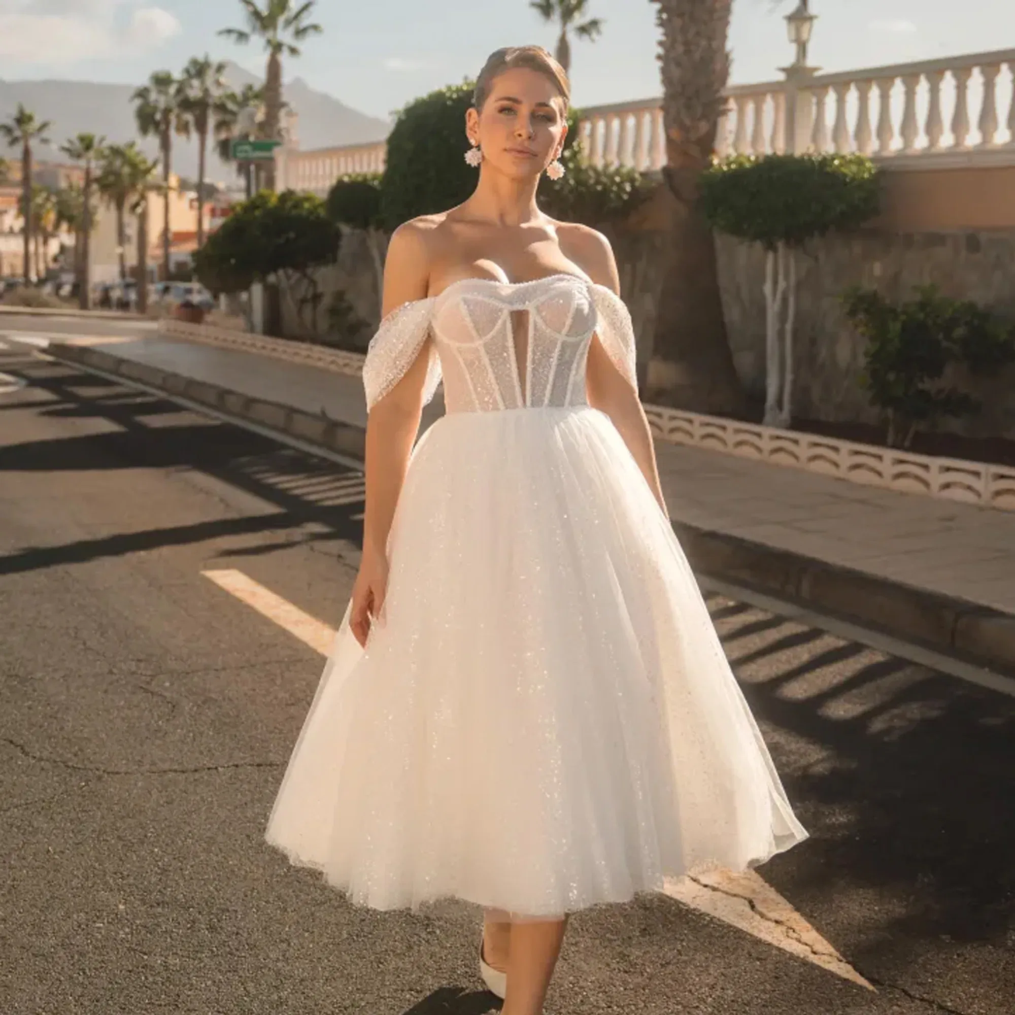 Junge Frau in einem romantischen, schimmernden Brautkleid mit ausgestelltem Tüllrock und freien Schultern posiert auf einer südlichen Straße im Abendlicht.