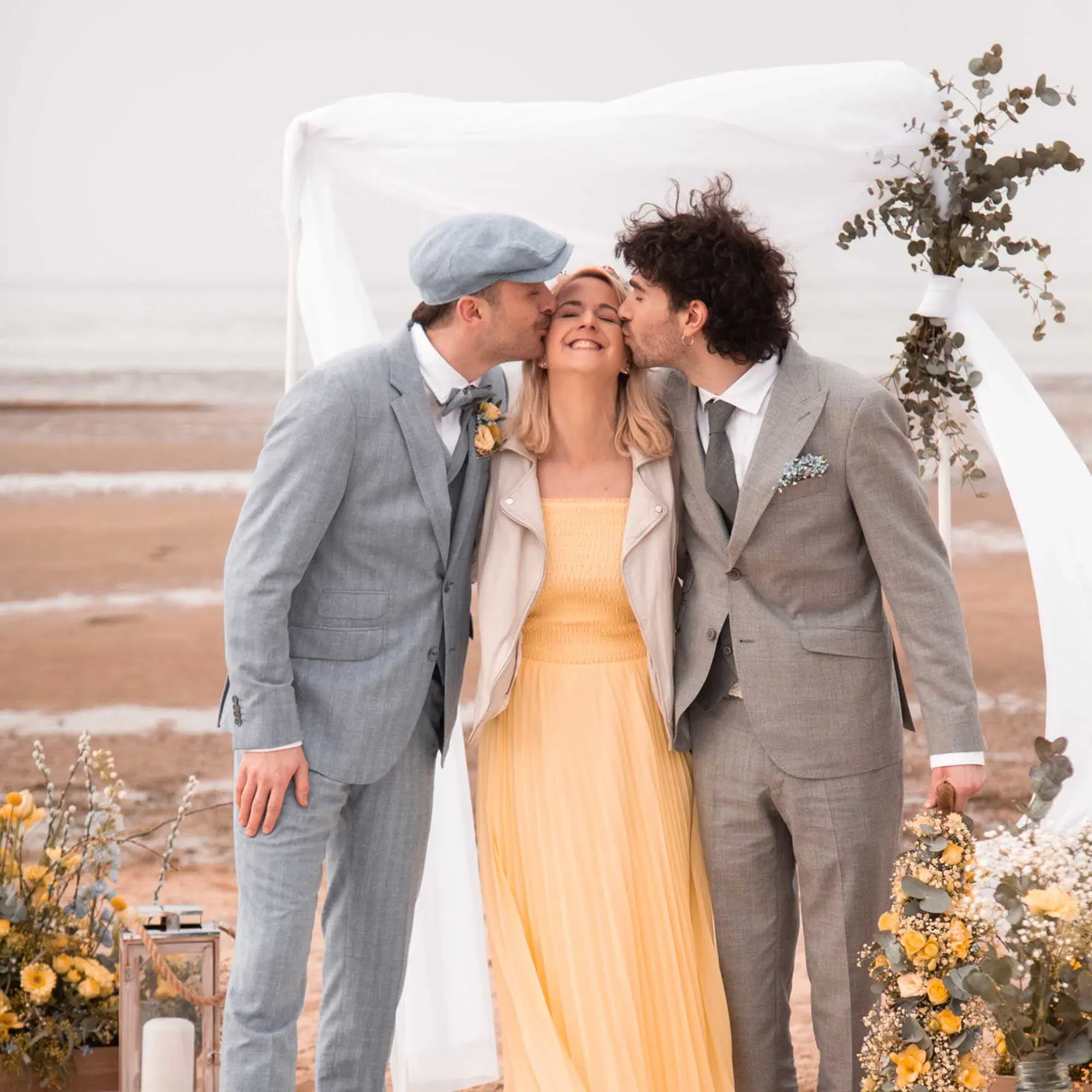 Fröhliche Szene bei einer LGBTQ+-Hochzeit am Strand – zwei festlich gekleidete Männer zeigen Zuneigung zu einer Frau unter einem weißen Baldachin mit Eukalyptus und gelben Blumen.