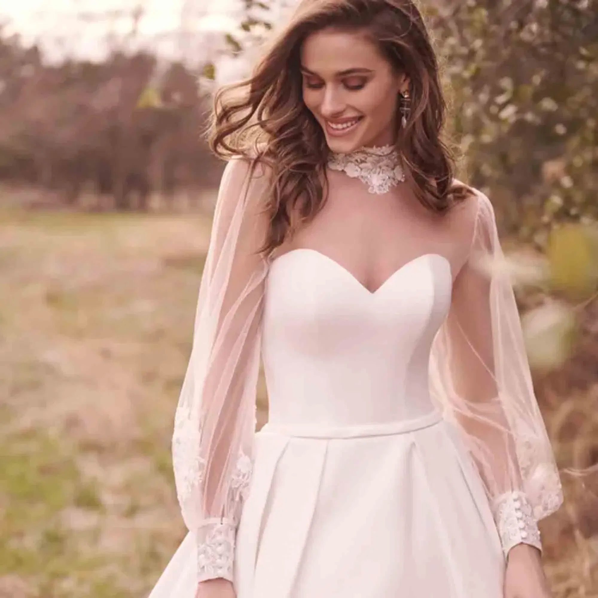 Zartes Hochzeitskleid mit Illusionsärmeln und feinem Spitzendetail am Hals – Braut lächelt verträumt in natürlicher Umgebung.