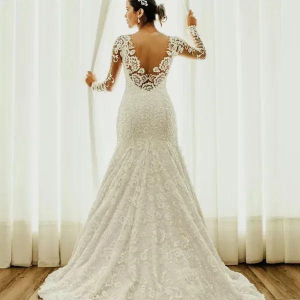 Braut in einem weißen, reich verzierten Hochzeitskleid mit tiefem Rückendekolleté und langen Ärmeln aus Spitze vor hellen Gardinen.