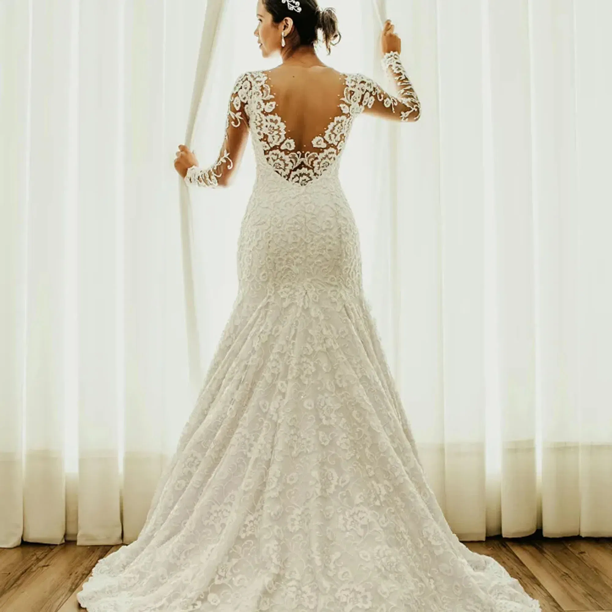 Braut in einem weißen, reich verzierten Hochzeitskleid mit tiefem Rückendekolleté und langen Ärmeln aus Spitze vor hellen Gardinen.