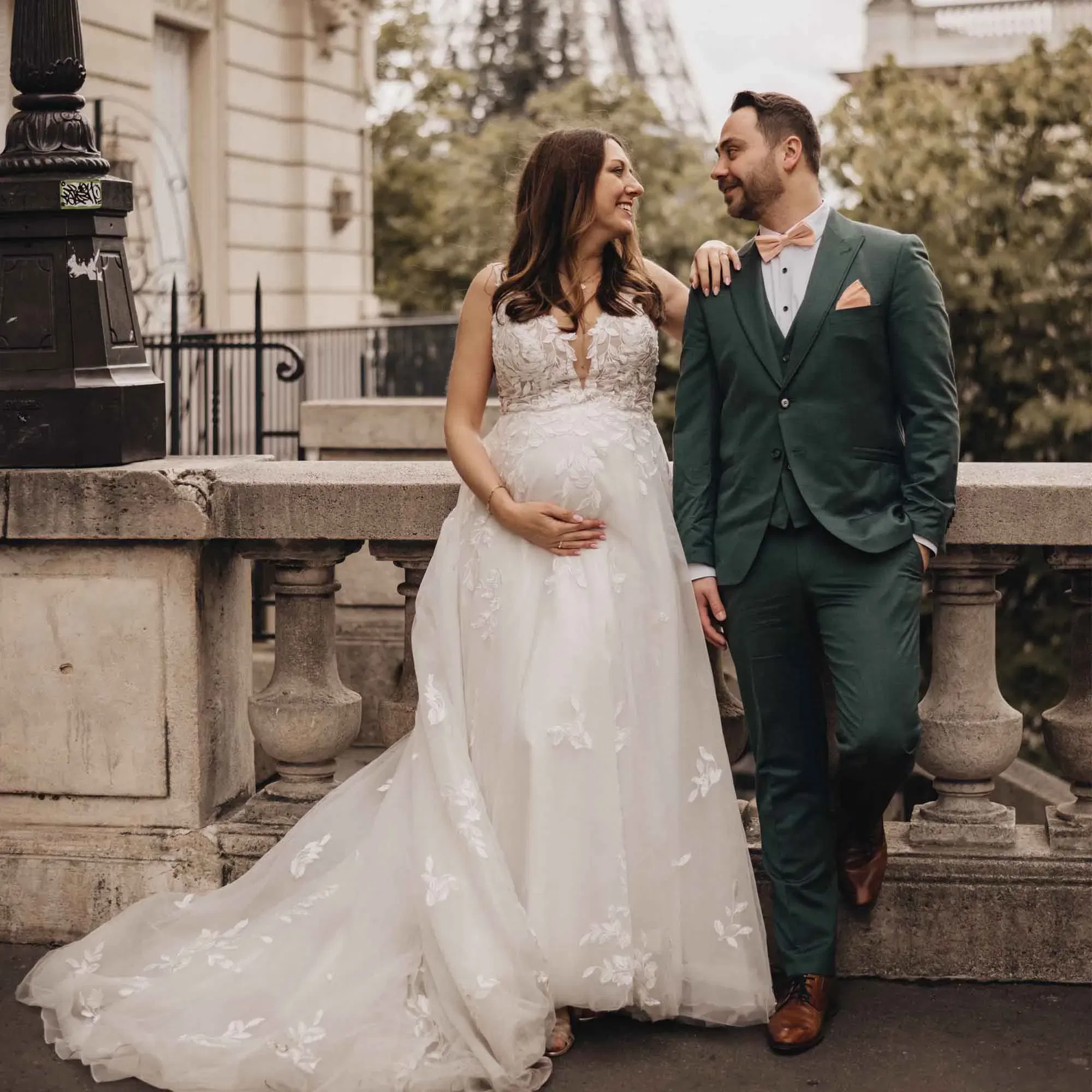Verliebtes Paar posiert für ein Babybauch-Fotoshooting in Paris: Sie im Brautkleid mit Schleier, er im grünen Hochzeitsanzug vor urbaner Kulisse.