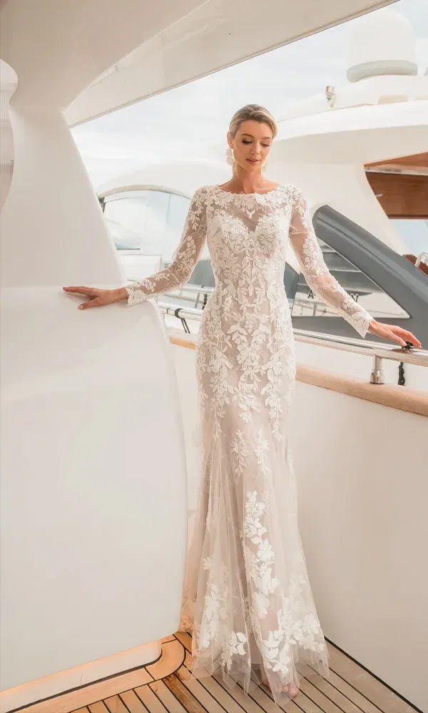 Elegante Braut in einem figurbetonten Brautkleid mit feinem Blütenmuster und Illusionsausschnitt, stehend auf einer stilvollen Yacht mit hellem Deck.