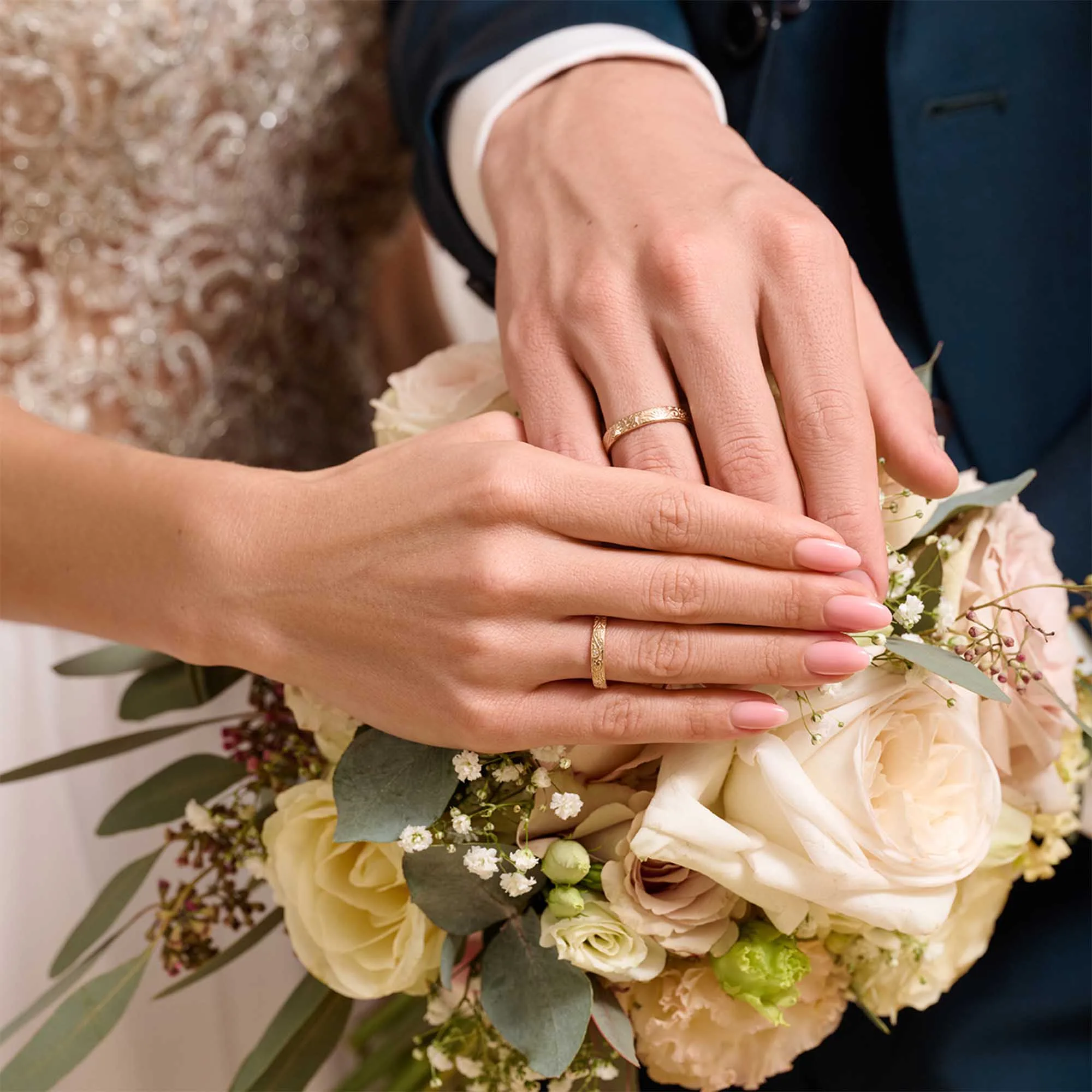Braut- und Bräutigam-Hände mit goldenen Eheringen liegen auf einem Brautstrauß mit cremefarbenen Rosen und Eukalyptusblättern.