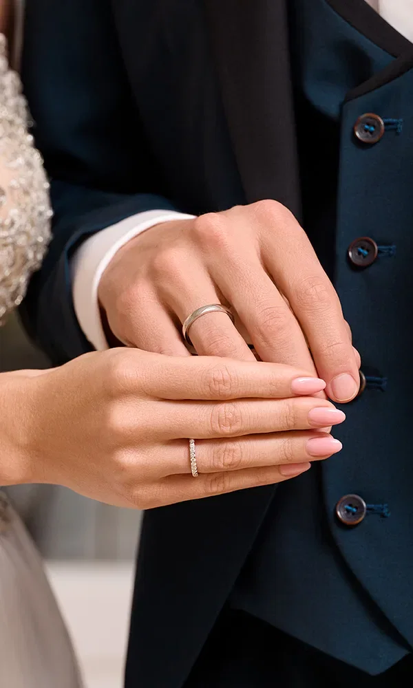 Nahaufnahme eines Brautpaars, das sich die Hände reicht – zu sehen sind ein schlichter Herrenring und ein Diamant-Ehering an der Hand der Braut.