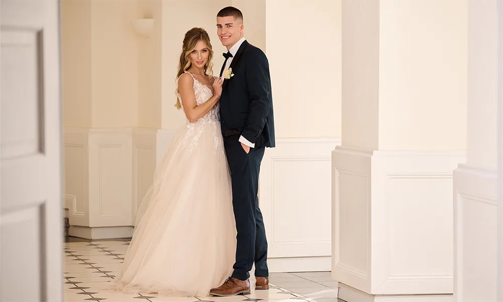 Brautpaar in eleganter Pose in einem hellen Flur – die Braut trägt ein cremefarbenes Kleid mit Spitzenoberteil, der Bräutigam einen dunkelblauen Anzug.