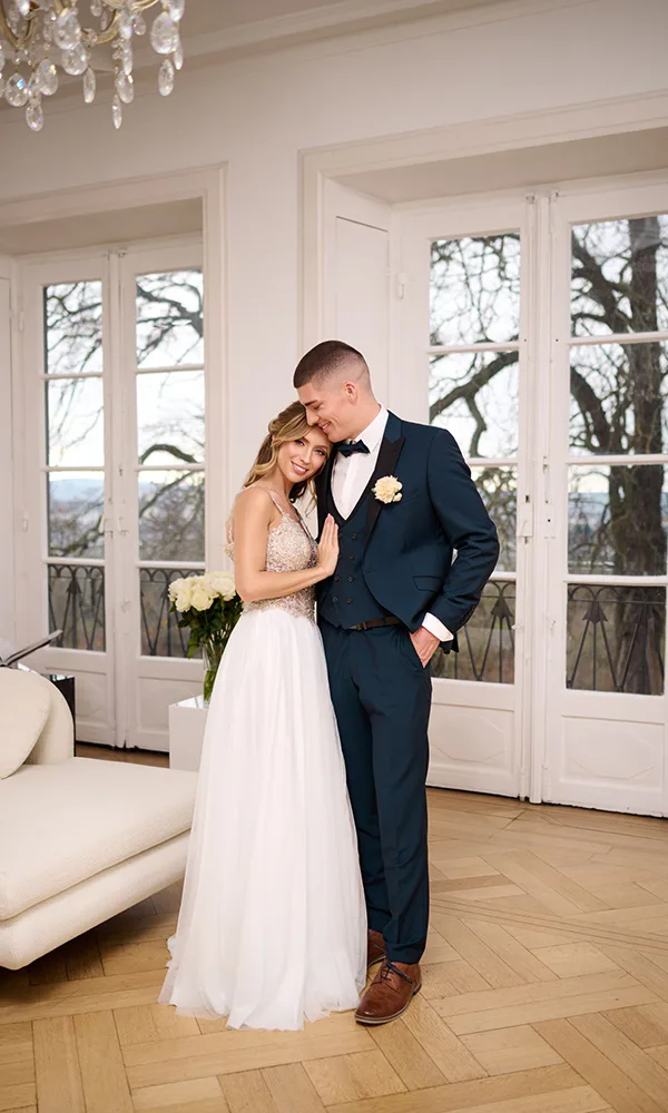 Zärtliche Umarmung eines stilvollen Brautpaars vor großen Fenstern – die Braut lehnt sich mit geschlossenen Augen an den Bräutigam.