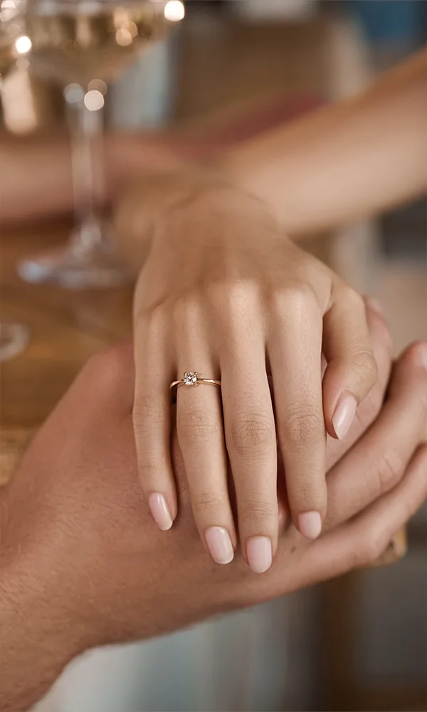 Close-up einer Hand mit zartem Solitär-Verlobungsring in Gold, die von einer zweiten Hand liebevoll gehalten wird.