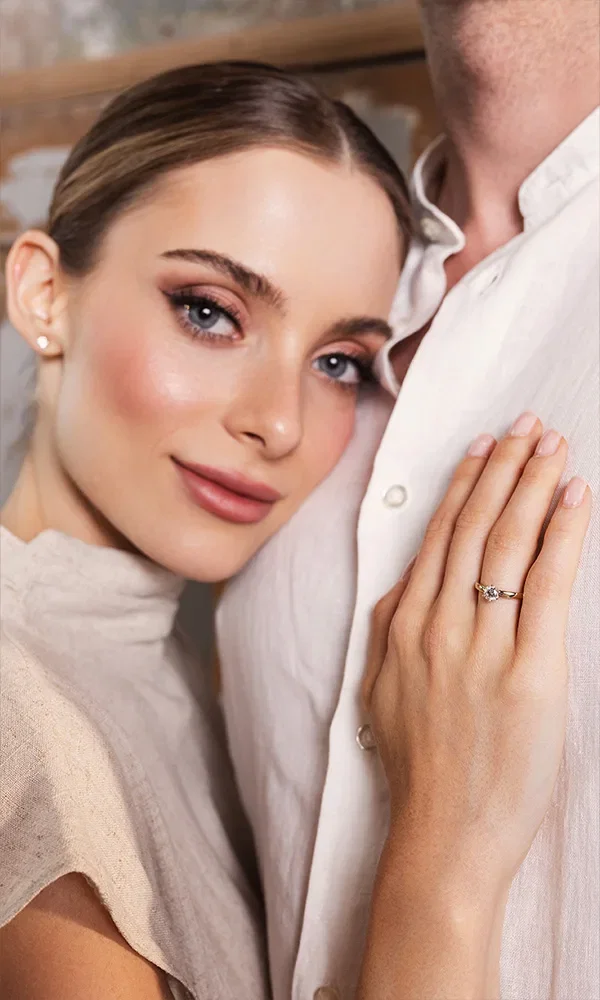 Porträt einer Frau mit elegantem Make-up, die ihre Hand mit einem funkelnden Verlobungsring an die Brust ihres Partners legt.