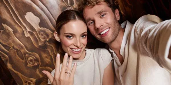 Ein Mann und eine Frau lächeln in die Kamera. Sie hält ihre Hand hoch. Daran ein Verlobungsring mit Diamant.