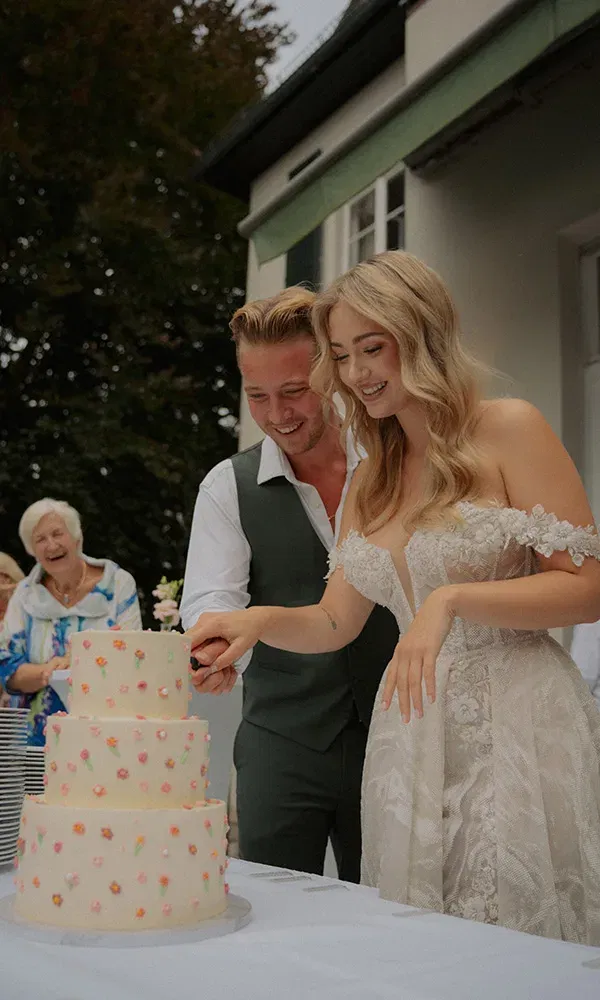 Paar lacht gemeinsam beim Anschneiden der Hochzeitstorte unter freiem Himmel.