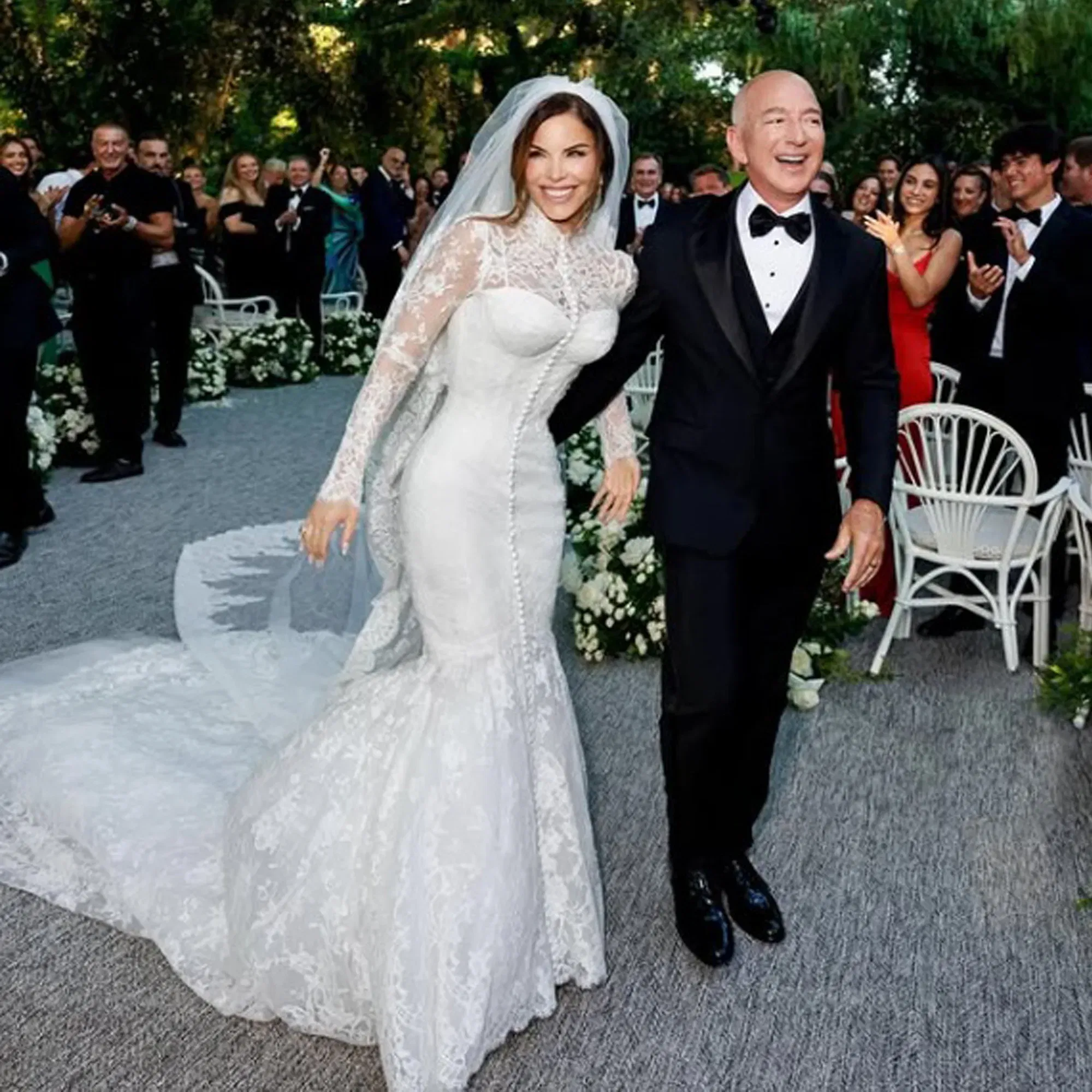 Frisch vermähltes Paar bei einer glamourösen Outdoor-Hochzeit, mit festlicher Dekoration aus weißen Blumen und Gästen in Abendgarderobe im Hintergrund.