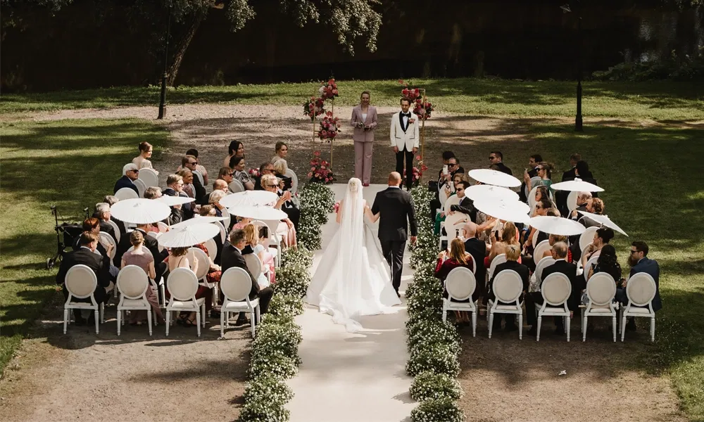 Braut und Vater gehen bei strahlendem Sonnenschein den geschmückten Gang entlang, umgeben von Gästen mit Sonnenschirmen.