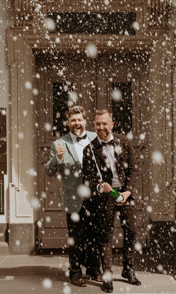Zwei Männer im Smoking feiern vor einer Tür, während aus der Flasche funkelnder Champagnerschaum sprüht.