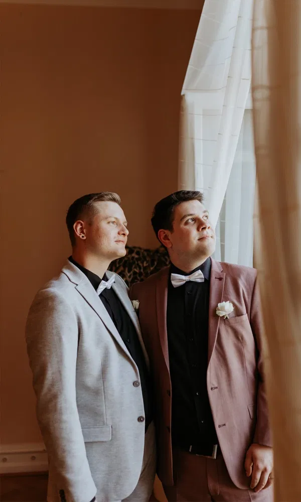 Zwei Männer in festlicher Kleidung genießen einen ruhigen Moment am Fenster und schauen in die Ferne.