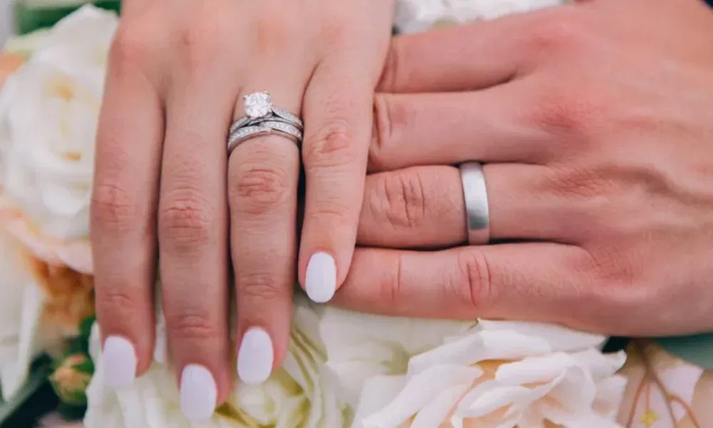 Hände eines Brautpaars mit Eheringen über einem Blumenstrauß.