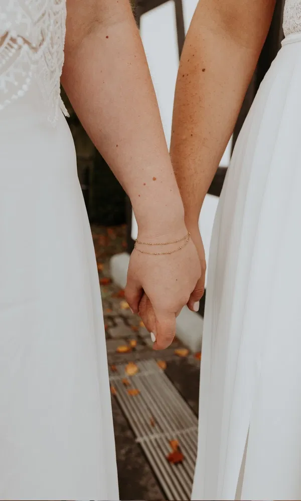 Zwei Bräute stehen nebeneinander und halten sich sanft an den Händen, Fokus auf dem Detail.