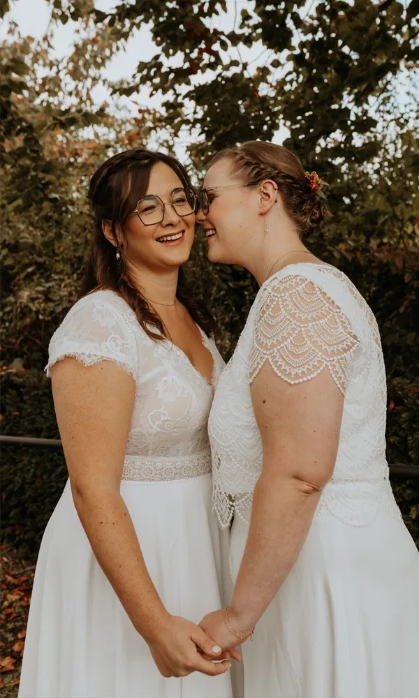 Zwei Bräute in weißen Kleidern teilen einen innigen Moment, während sie sich gegenseitig zulächeln.